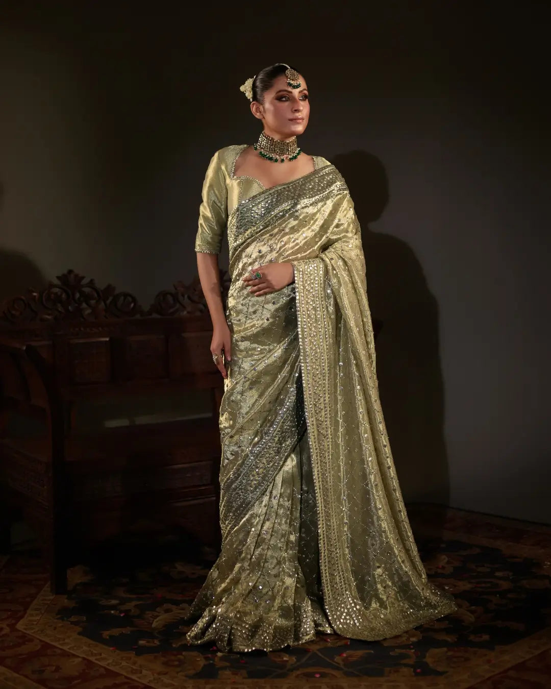 Woman in a gold embroidered saree with traditional jewelry, posing indoors on a dark rug.