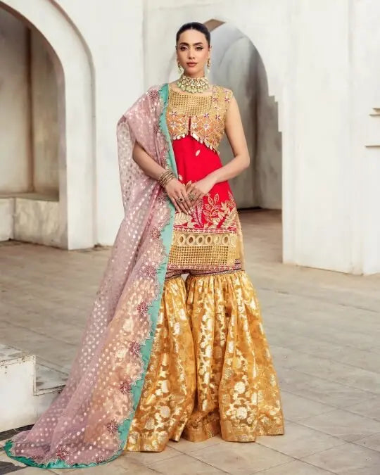 Woman in luxury Pakistani formal dress with gold sharara, red embroidered kameez, and net dupatta