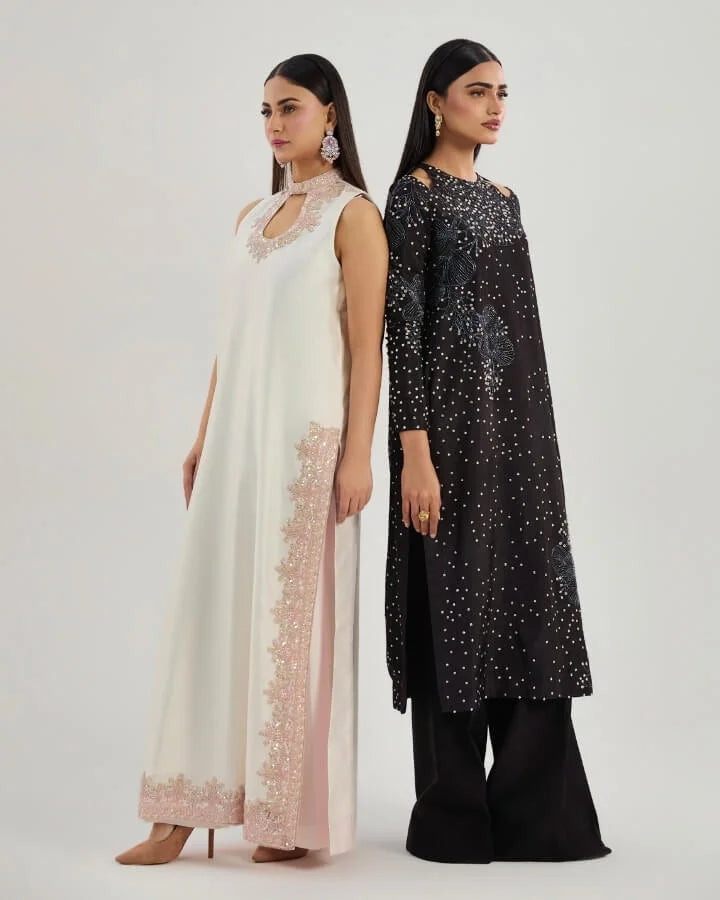 Two women in elegant formal Pakistani dresses, one in white with pink embroidery and one in black with silver embellishments, standing against a neutral background.
