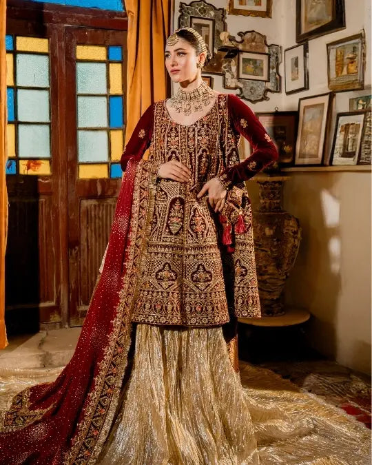 Woman in a traditional maroon and gold bridal dress with intricate embroidery, standing indoors.