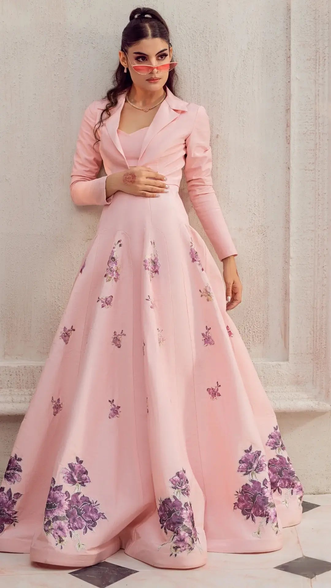 Woman in a blush pink formal gown with purple floral embroidery, standing against a wall