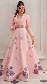 Woman in a blush pink formal gown with purple floral embroidery, standing against a wall