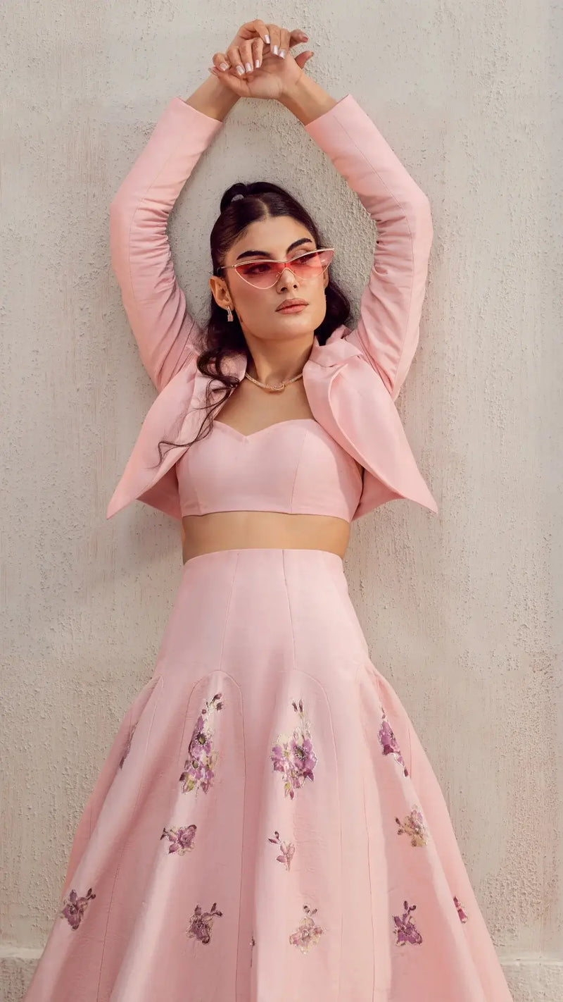 Woman in pastel pink crop top and jacket with floral embroidered maxi skirt, posing against wall
