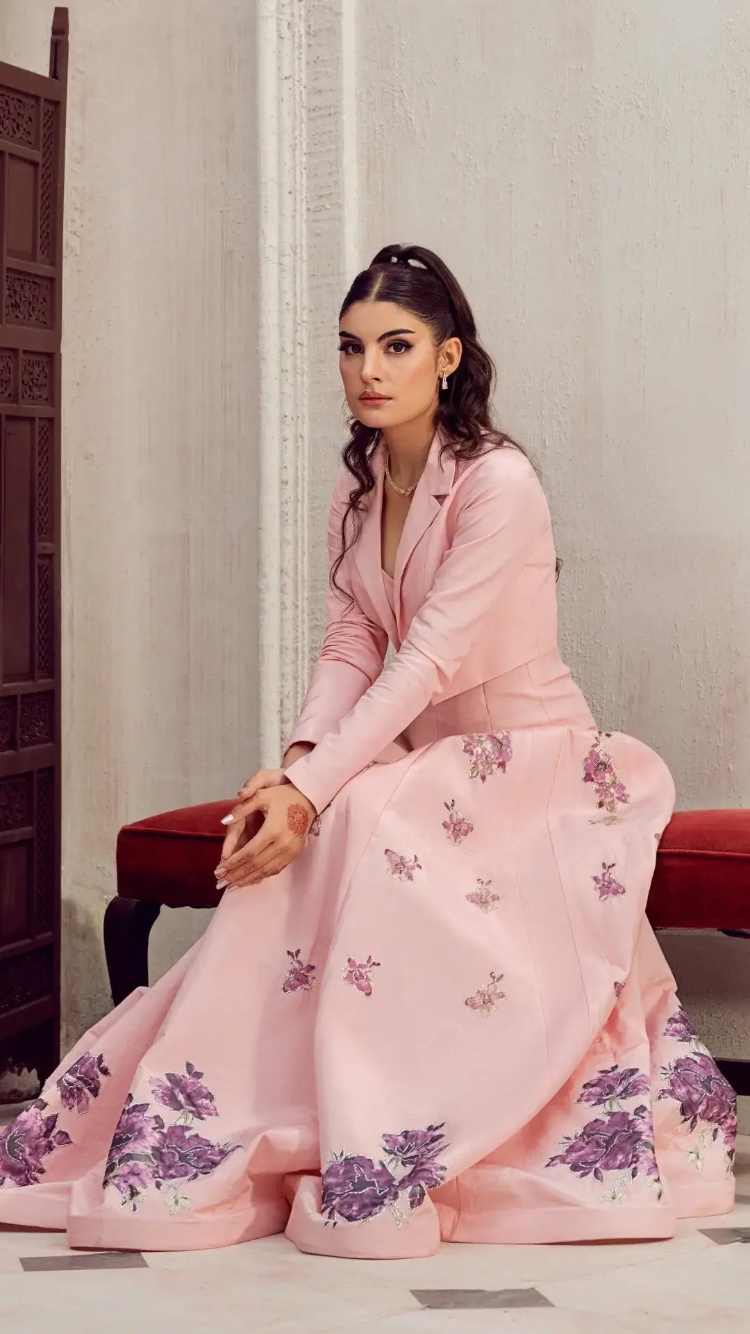 Woman in elegant pastel pink formal dress with floral embroidery, seated indoors on a red bench