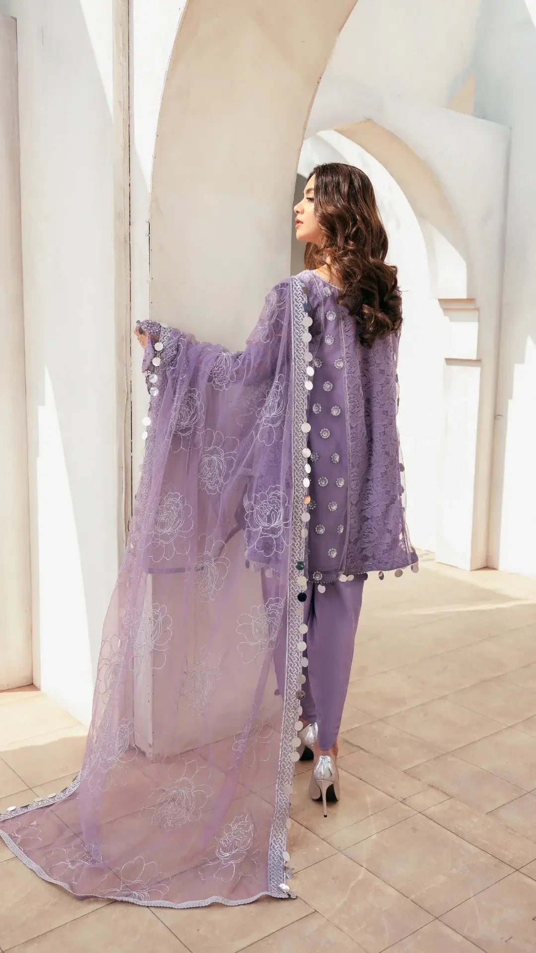 Woman in a lavender traditional outfit with intricate designs in a bright, architectural setting.