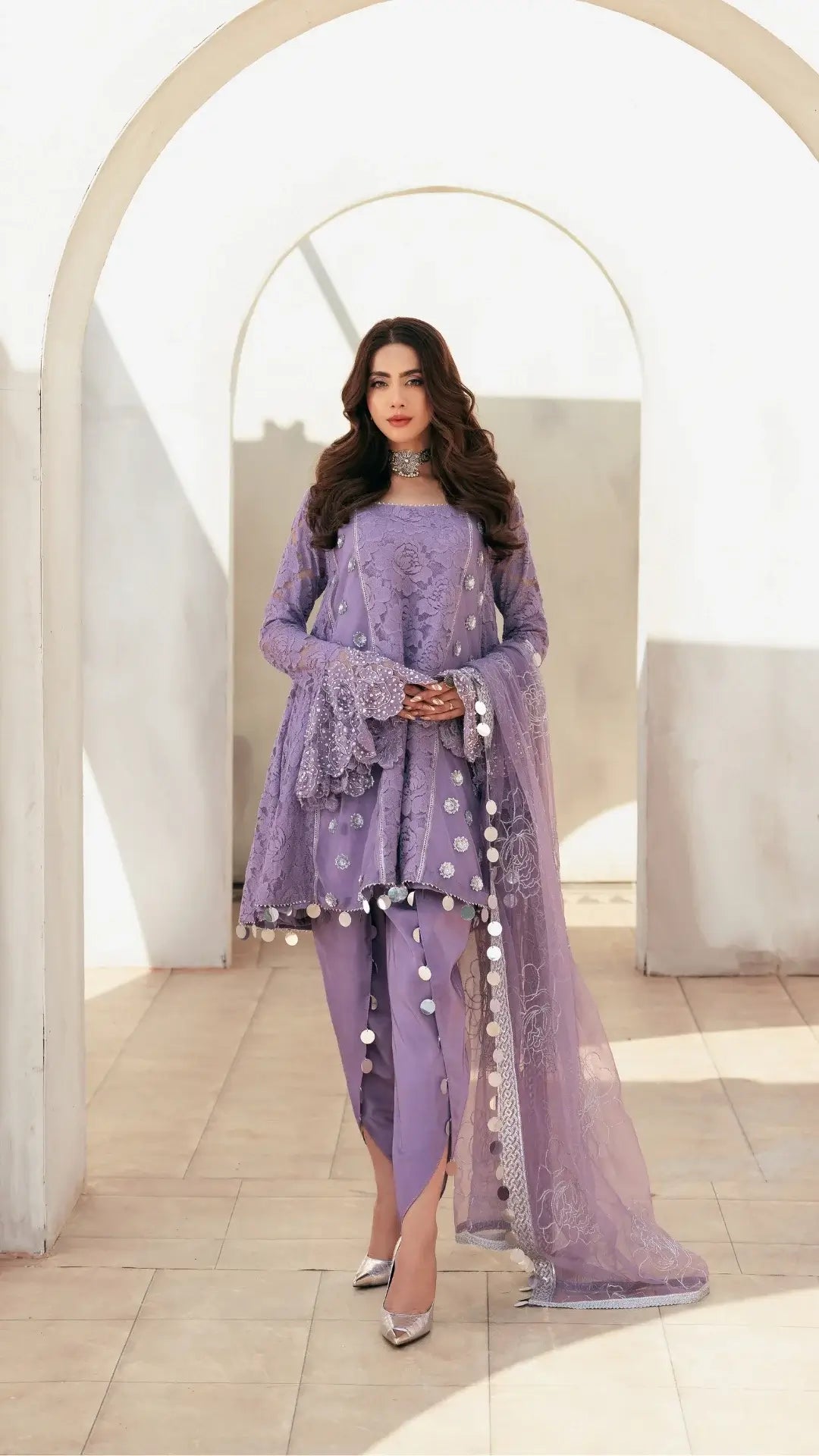 Woman wearing a lavender traditional outfit with intricate designs in a bright interior setting.