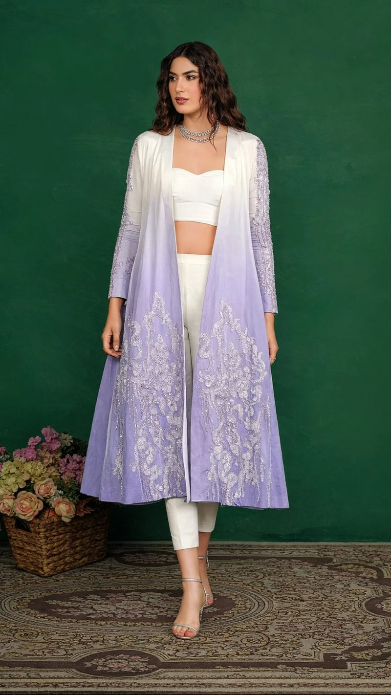 Woman wearing a long light purple embroidered jacket over a white outfit against a green background