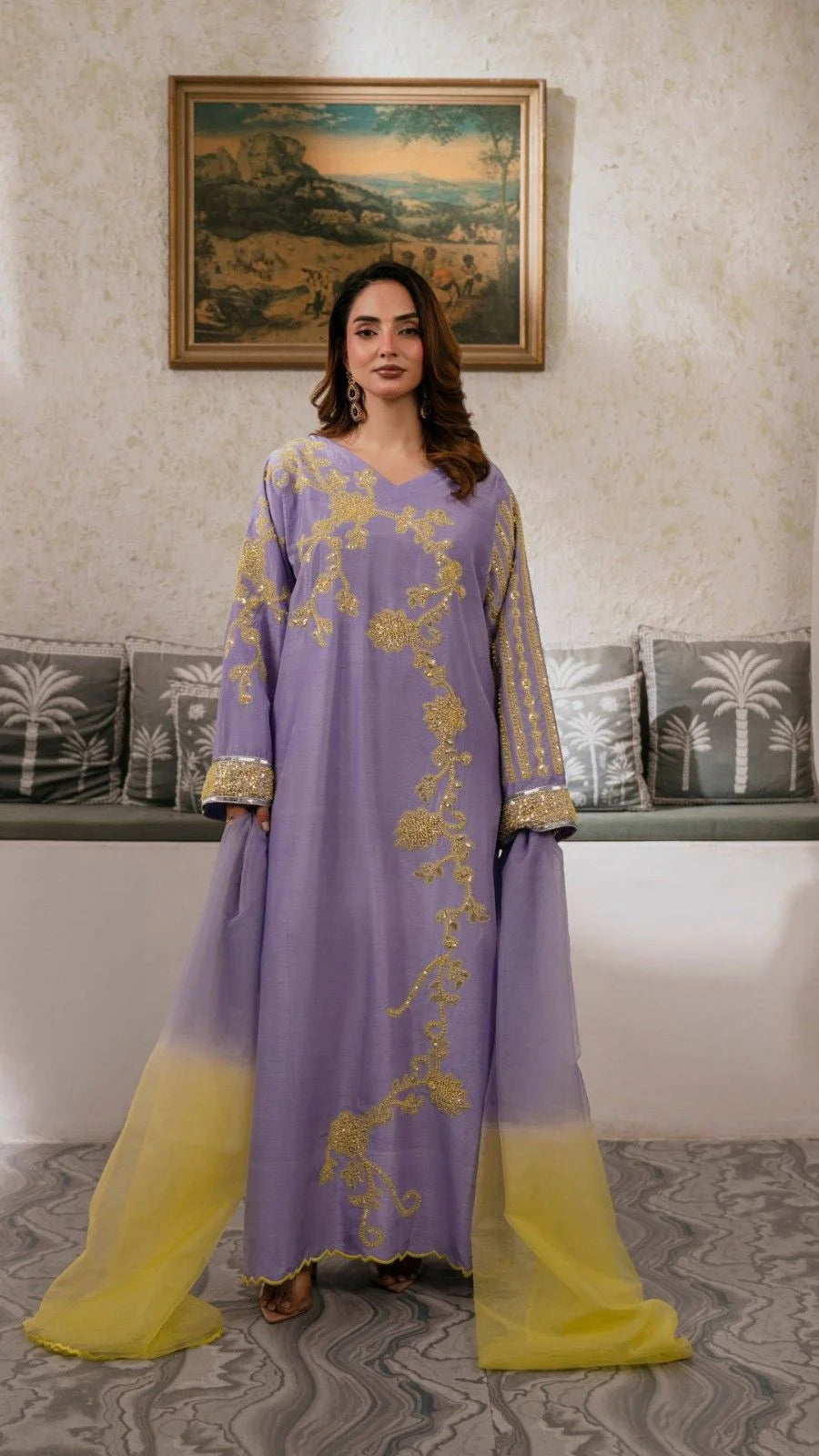 Woman wearing a lavender outfit with gold embroidery in a room with decorative pillows and a painting.