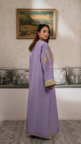 Woman in a long, light purple dress with gold embroidery standing in a room with decorative cushions and a painting on the wall.