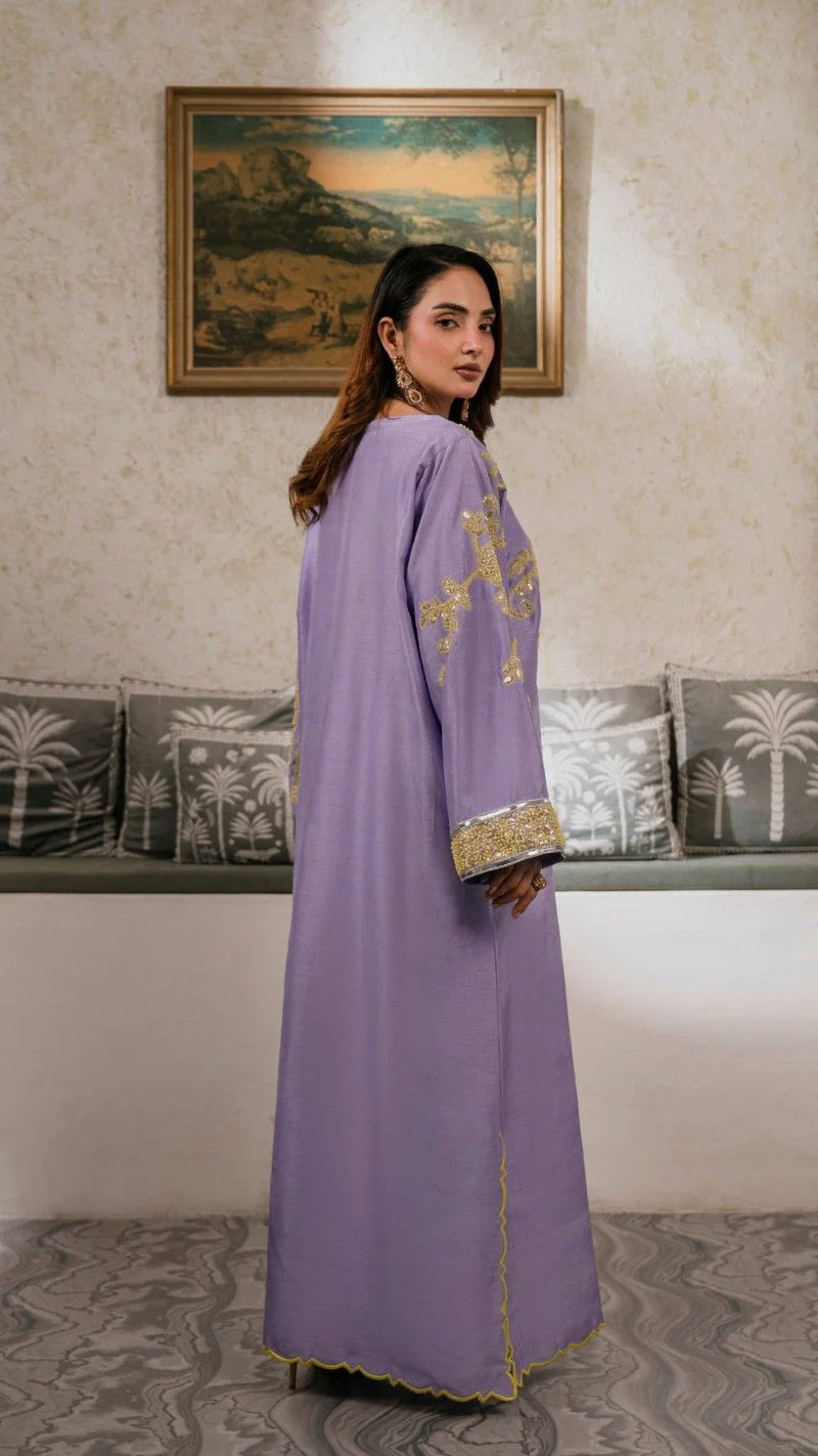 Woman in a long, light purple dress with gold embroidery standing in a room with decorative cushions and a painting on the wall.