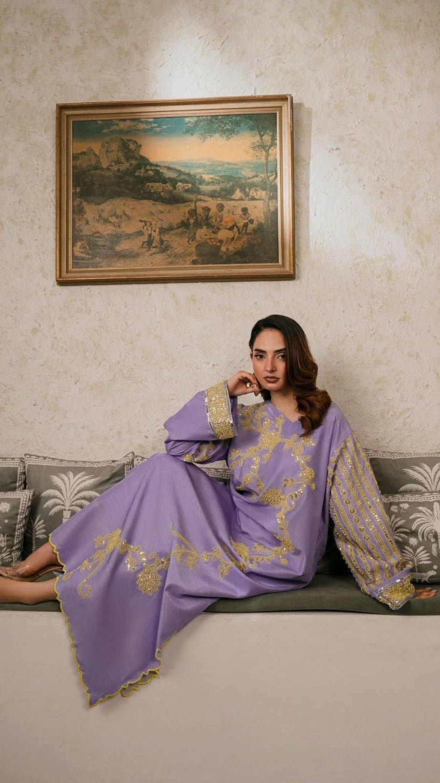 Woman in a purple dress with gold embroidery sitting on a couch in a room with a painting on the wall.
