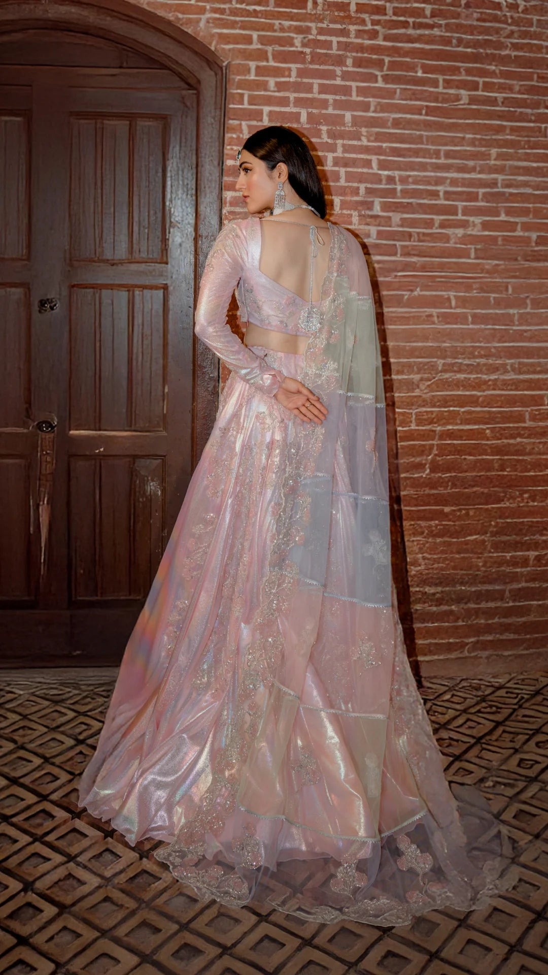 Woman in pastel pink embroidered formal lehenga with dupatta, standing by brick wall