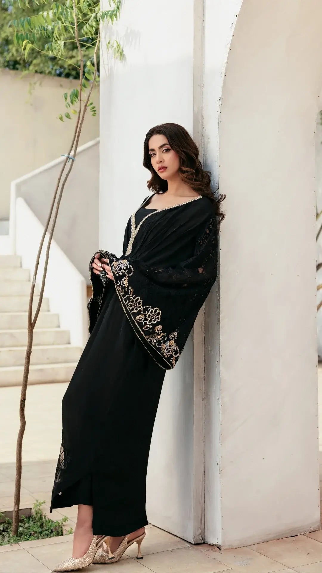 Woman in a black dress with gold embroidery standing against a white column.