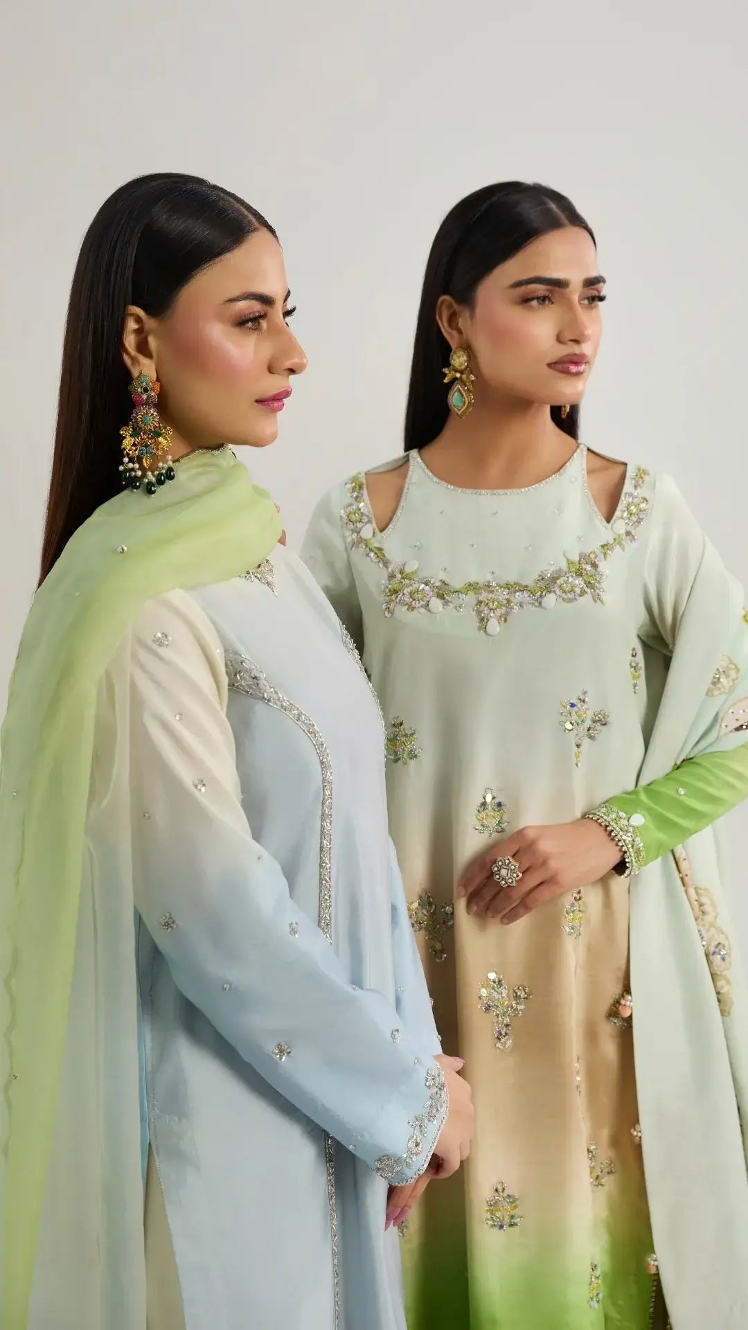 Two women wearing light-colored traditional outfits with floral embroidery on a plain background