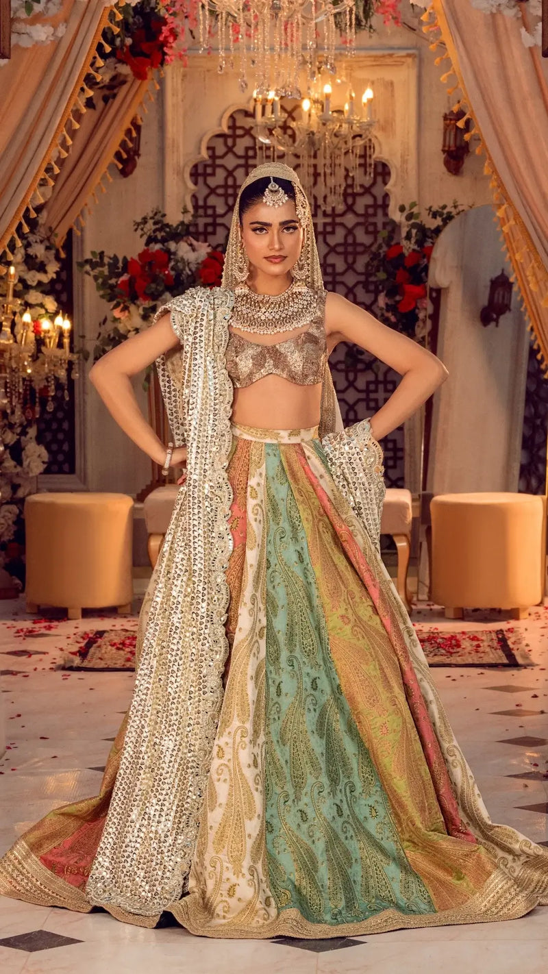 Woman in luxury Pakistani bridal lehenga with gold crop top, multicolor embroidered skirt, and dupatta, standing in ornate indoor setting with chandeliers and flowers.