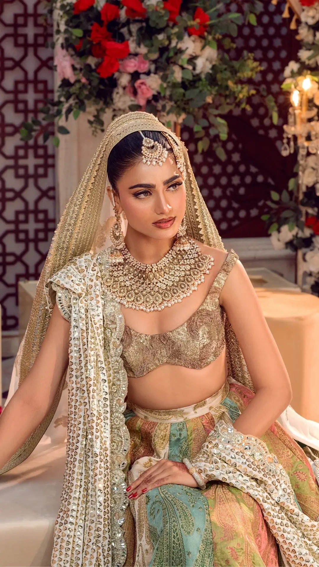 Woman in a gold embroidered bridal lehenga with ornate jewelry, seated indoors with floral decor