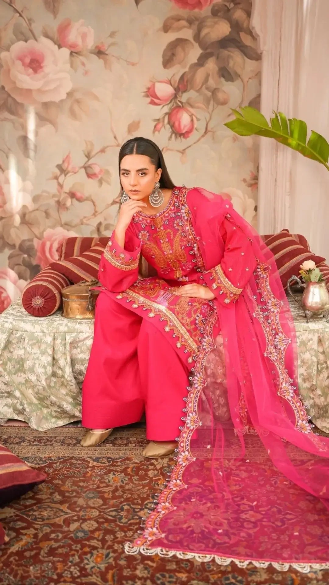 Woman in a traditional pink outfit with intricate designs sitting in a decorated room.