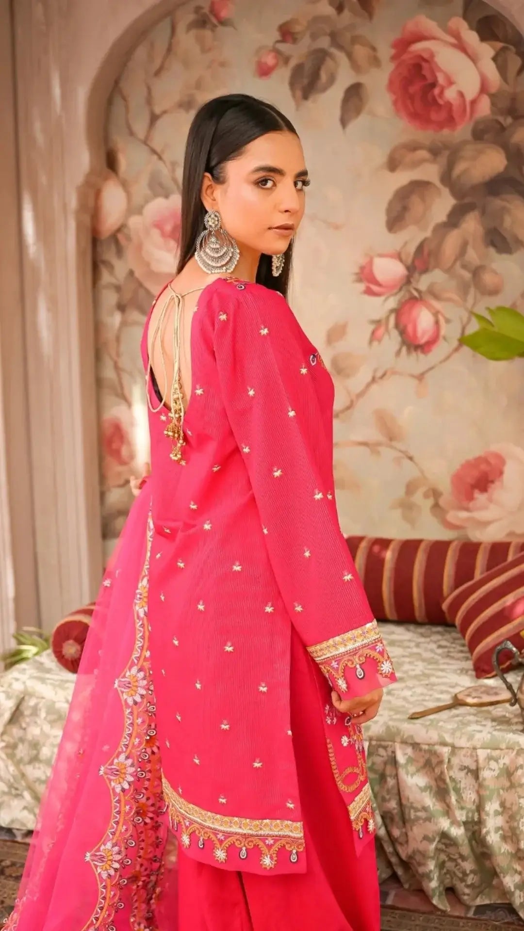 Woman in a bright pink traditional outfit with floral patterns in a decorated room.