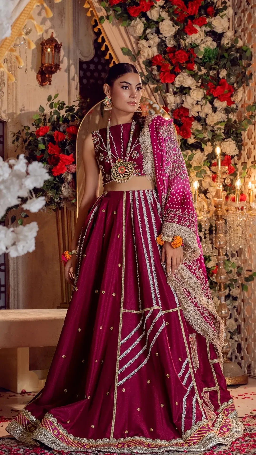 Woman in fuchsia Pakistani formal dress with gold embroidery, floral dupatta, and jewelry, standing in elegant, flower-adorned setting.