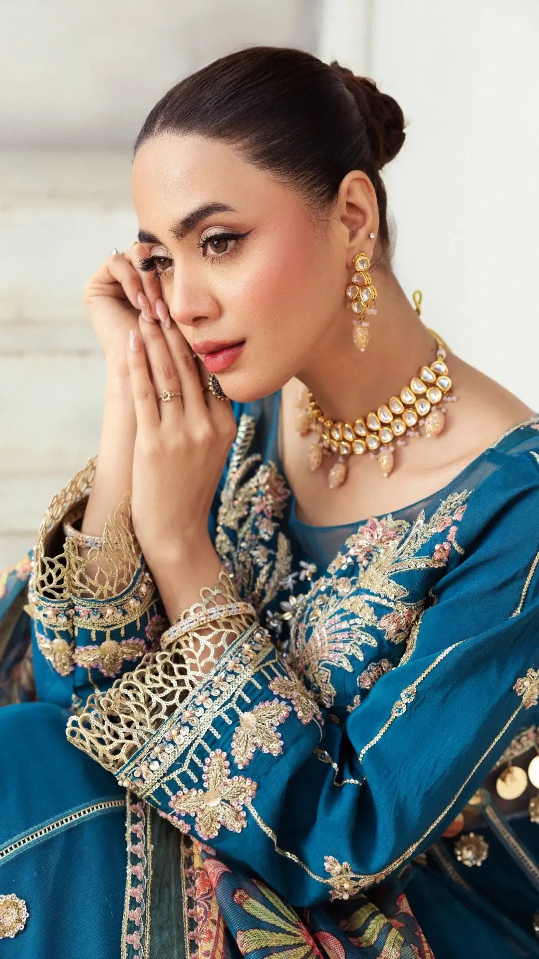 Woman in traditional blue embroidered outfit with gold jewelry against a neutral background
