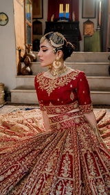 Woman in red and gold heavily embroidered bridal lehenga with traditional jewelry, seated indoors