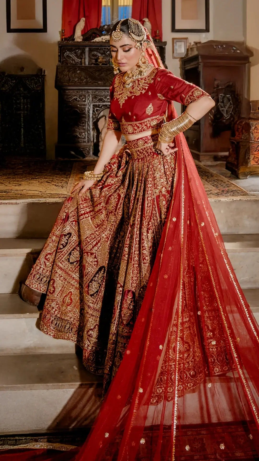 Woman in ornate red and gold Pakistani bridal dress with intricate embroidery, dupatta, and jewelry