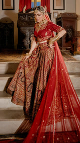 Woman in ornate red and gold Pakistani bridal dress with intricate embroidery, dupatta, and jewelry
