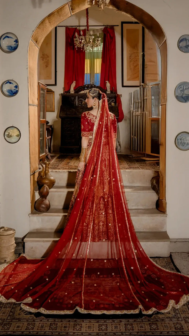 Woman in elegant red bridal dress with gold embroidery and long dupatta, traditional Pakistani formal wear, standing indoors.