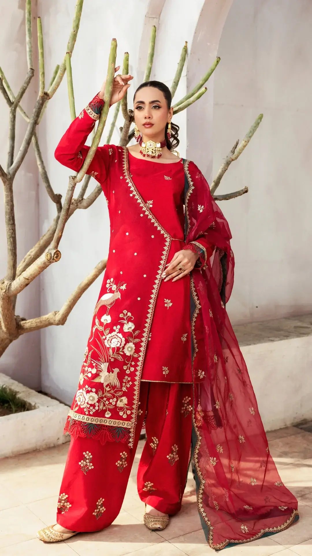 Woman in a red embroidered traditional outfit standing against a neutral wall with branches.