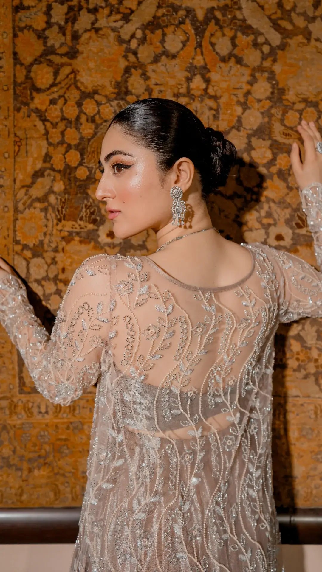 Woman in an elegant dress with intricate patterns against a textured wall.