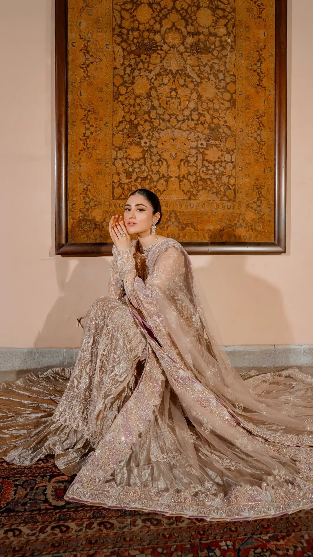 Woman in an elegant beige embroidered outfit sitting on a patterned rug with a decorative wall hanging in the background.