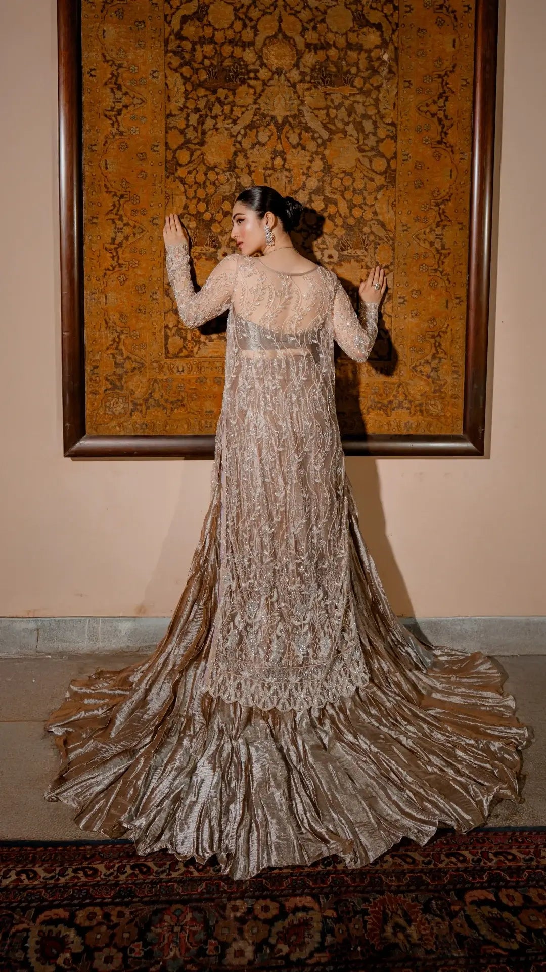 Woman in a long ivory embroidered formal dress with train, standing indoors facing ornate tapestry.