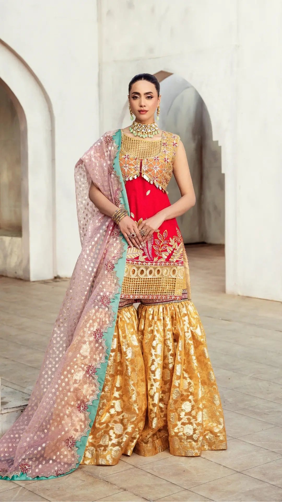 Woman in traditional outfit with red blouse and gold saree standing in a decorative setting.