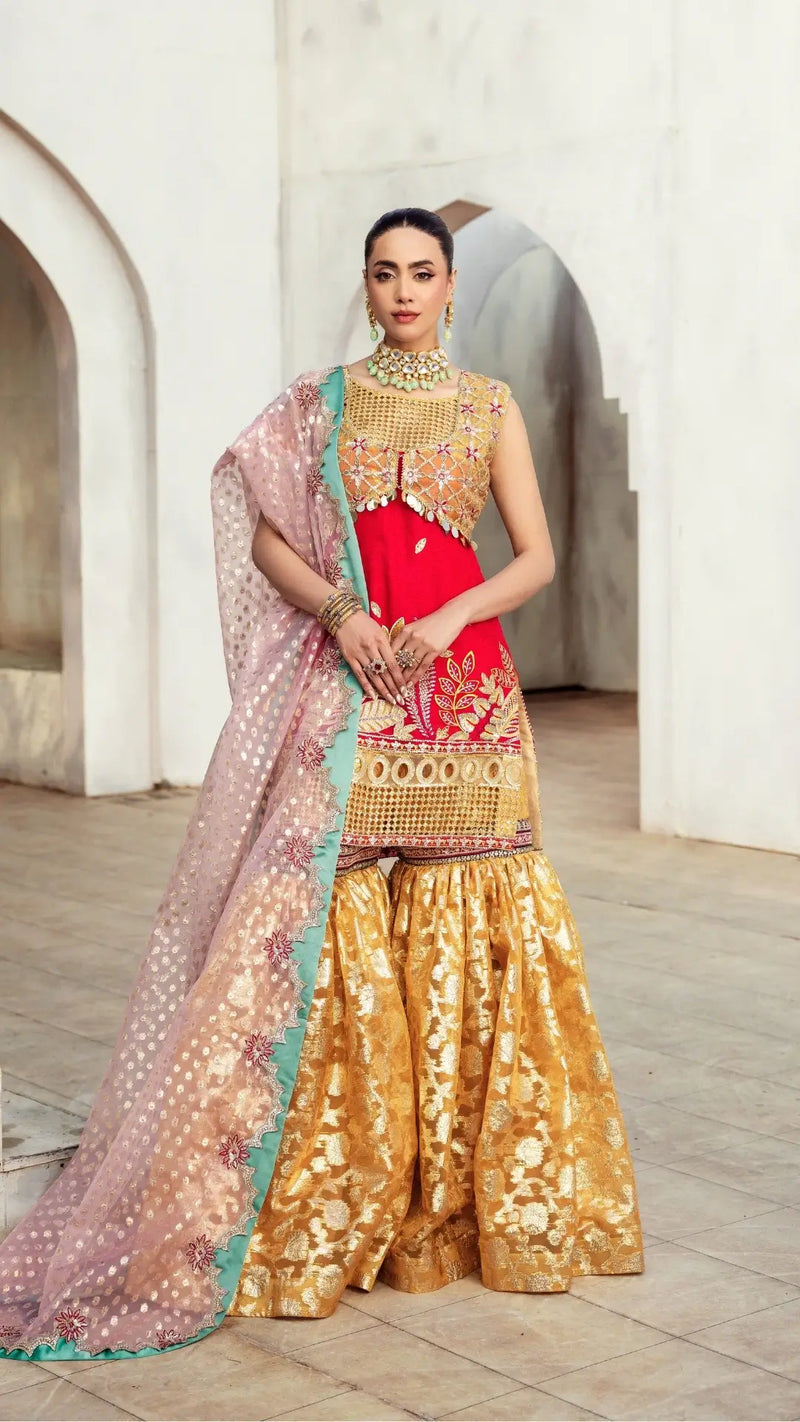 Woman in traditional outfit with red blouse and gold saree standing in a decorative setting.