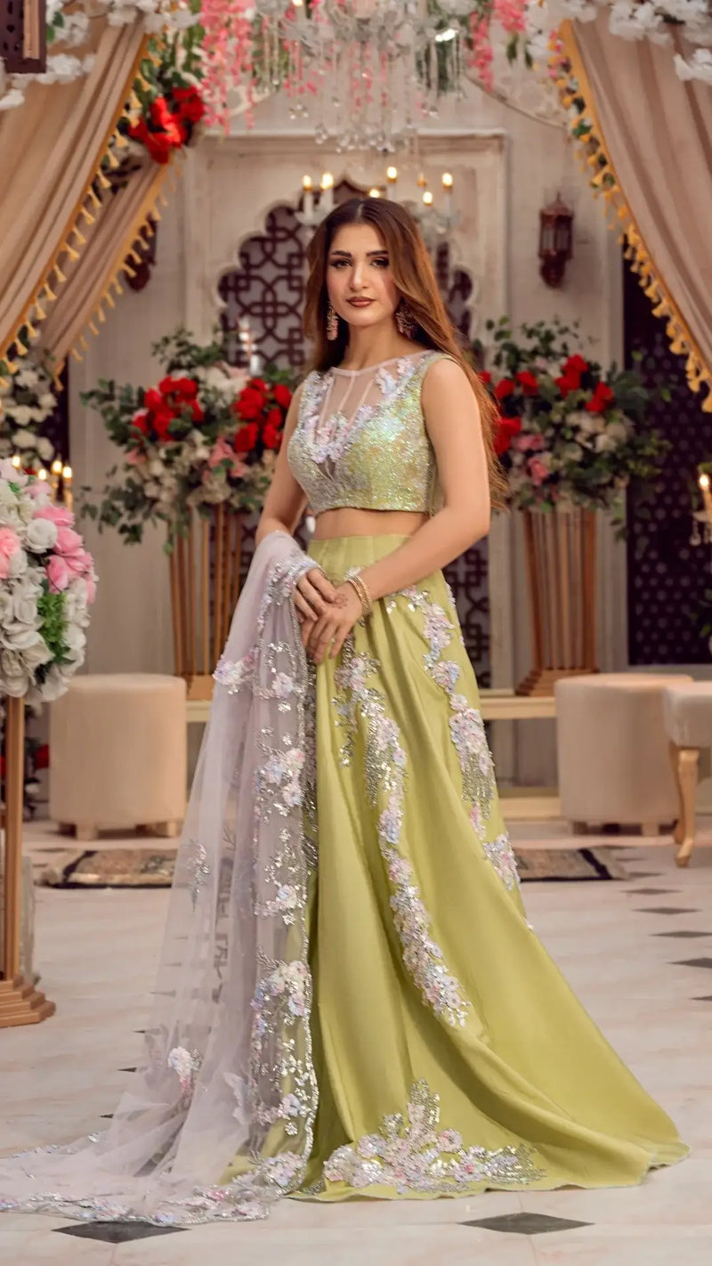 Woman in a green embellished formal lehenga dress with floral embroidery, holding dupatta, in a decorated indoor setting with flowers and chandeliers.