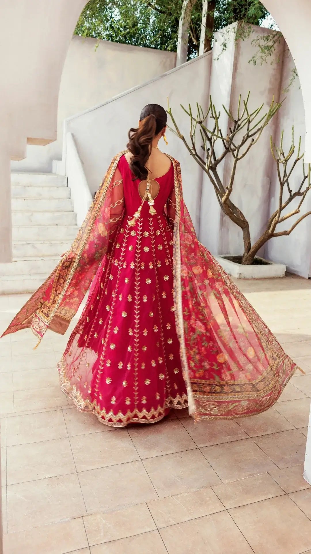 Woman in a red embroidered traditional outfit standing in a well-lit room with a plant in the background.