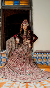 Woman in maroon heavily embroidered lehenga set with traditional jewelry, seated by ornate window