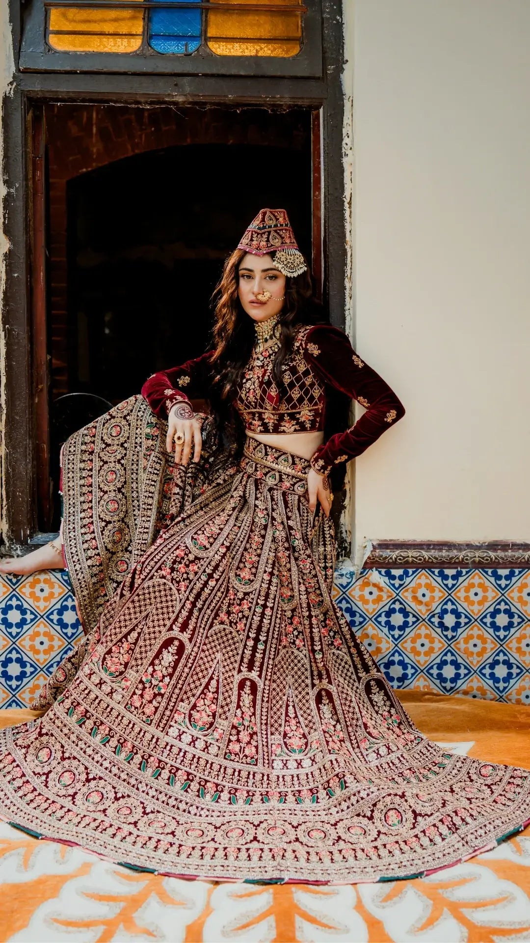 Woman in traditional embroidered outfit with a decorative headpiece, standing against a tiled wall.