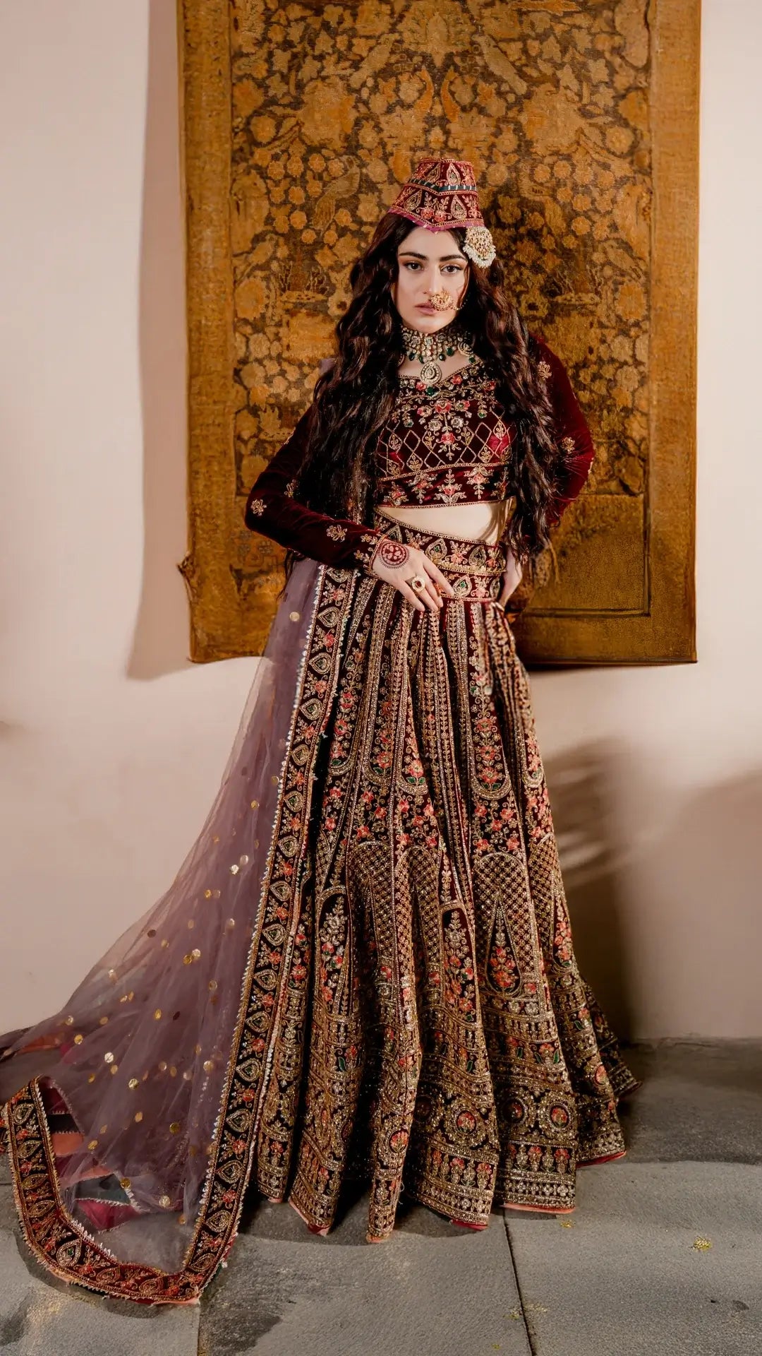 Woman in traditional embroidered outfit with a decorative headpiece against a textured wall.