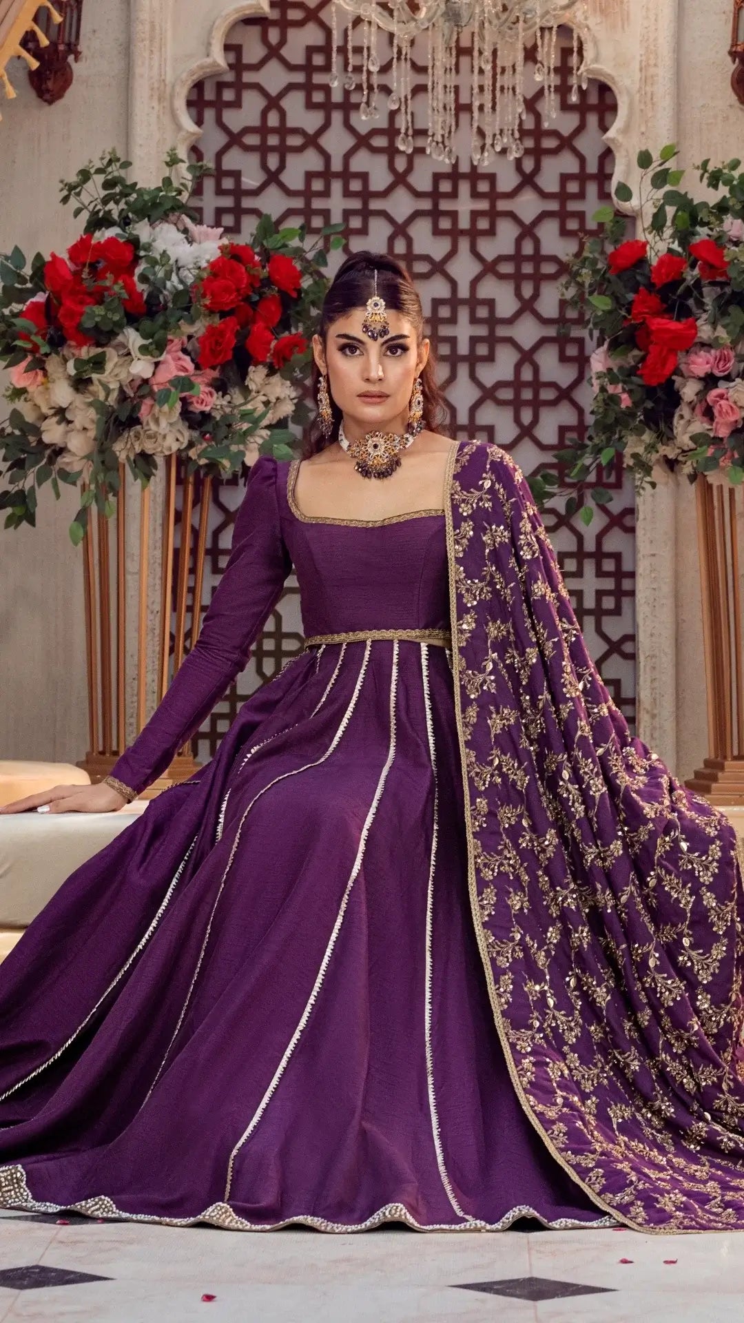 Woman wearing a purple formal Pakistani dress with gold embroidery, seated in an elegant floral setting.