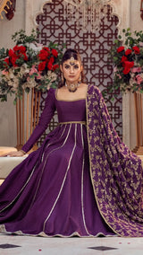 Woman wearing a purple formal Pakistani dress with gold embroidery, seated in an elegant floral setting.