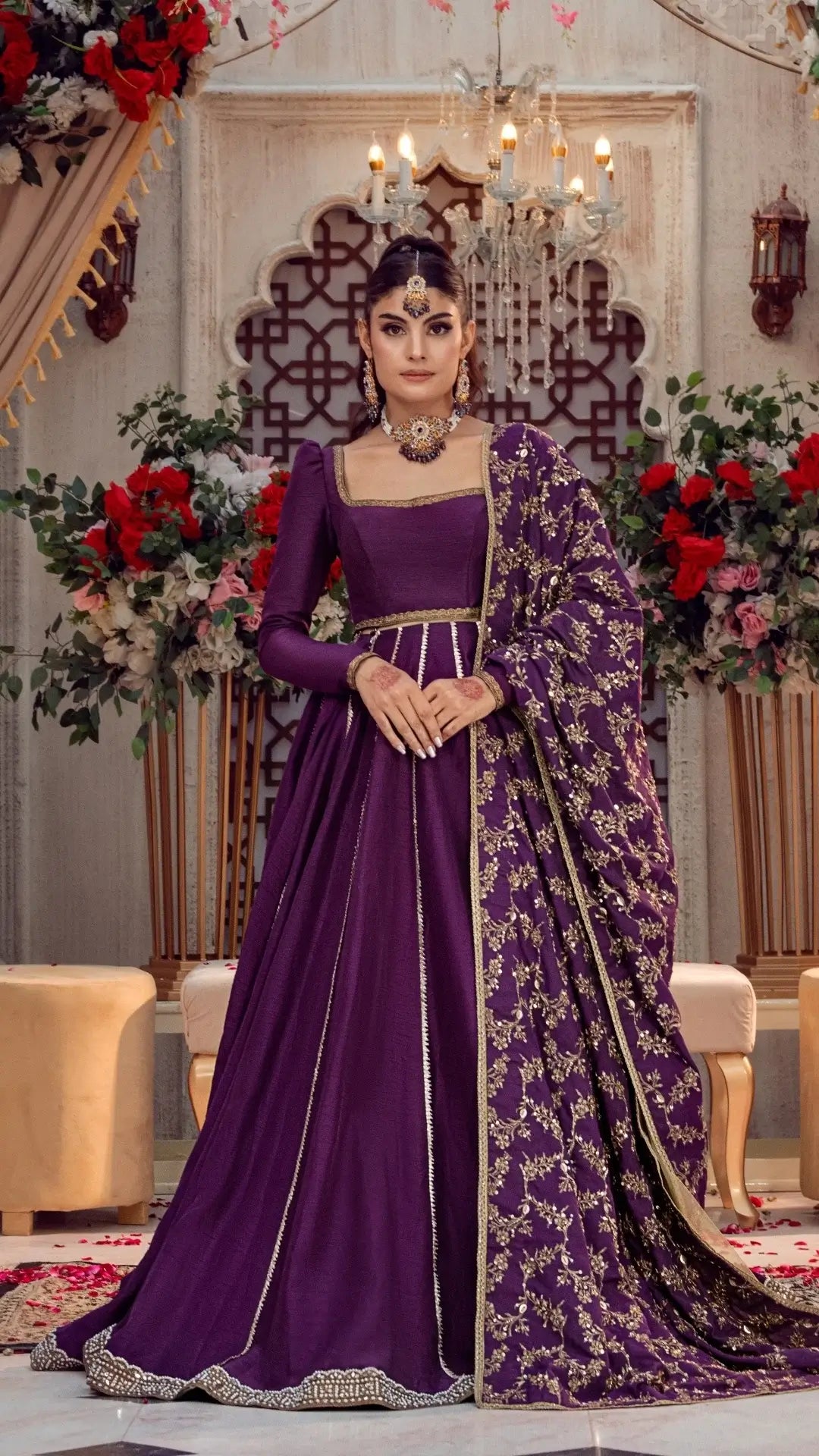 Woman in luxurious purple formal Pakistani dress with gold embroidery, standing in elegant indoor setting with floral décor.