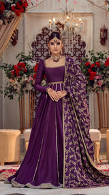 Woman in luxurious purple formal Pakistani dress with gold embroidery, standing in elegant indoor setting with floral décor.
