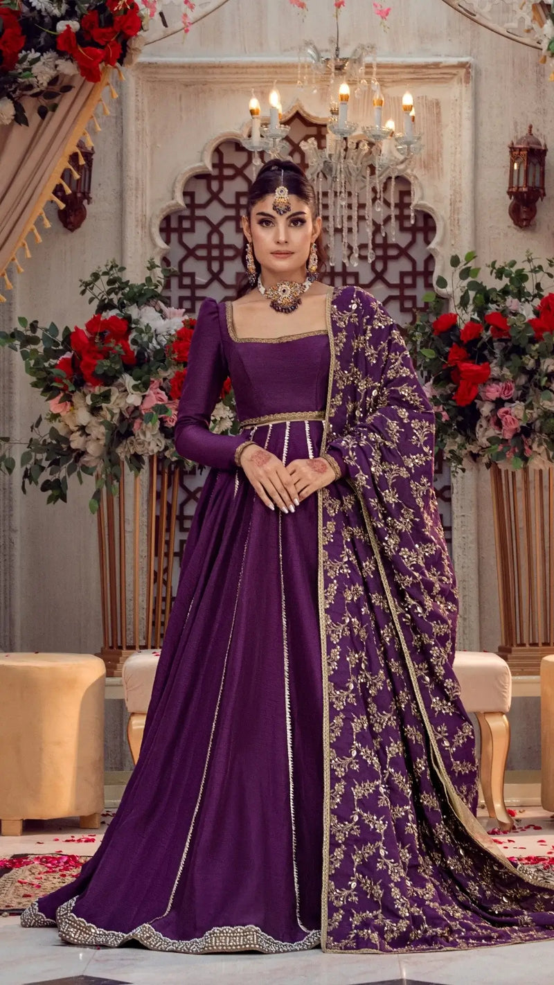 Woman in luxurious purple formal Pakistani dress with gold embroidery, standing in elegant indoor setting with floral décor.