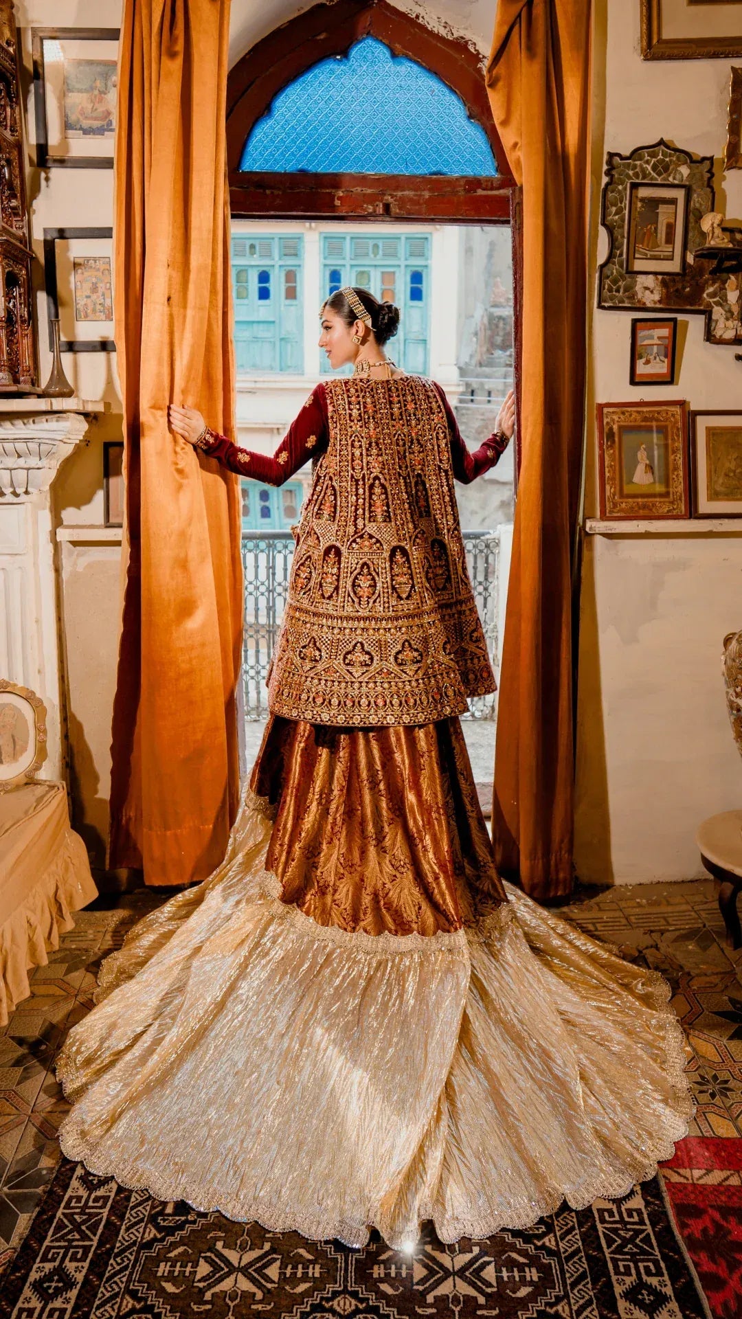 Woman in traditional embroidered outfit standing in a decorated room with orange curtains and decorative items.