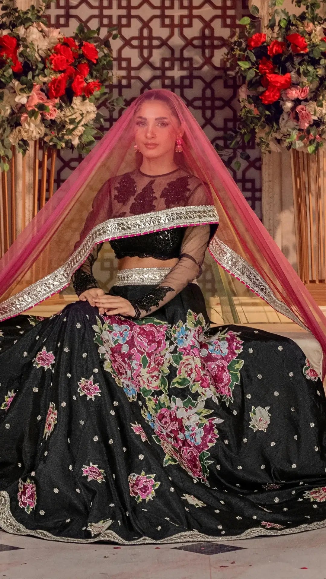 Woman wearing black floral embroidered Pakistani formal dress with pink dupatta, sitting in decorated setting with flowers.