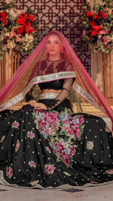 Woman wearing black floral embroidered Pakistani formal dress with pink dupatta, sitting in decorated setting with flowers.