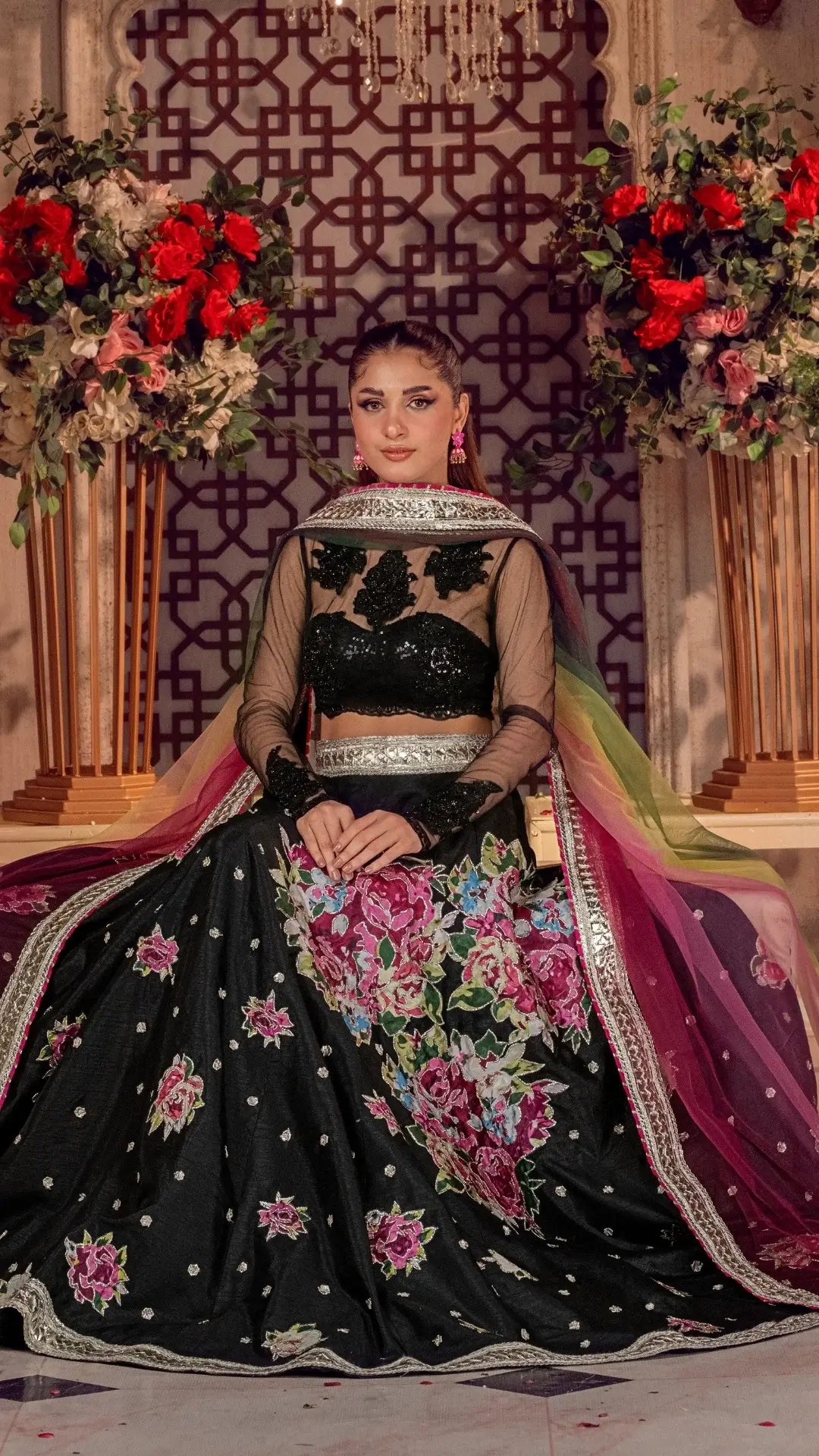 Woman wearing luxury black Pakistani formal dress with floral embroidery and dupatta, seated amid floral arrangements.