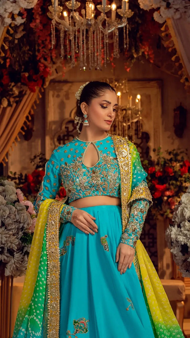 Woman in an embroidered turquoise formal dress with dupatta, posing at a floral indoor event