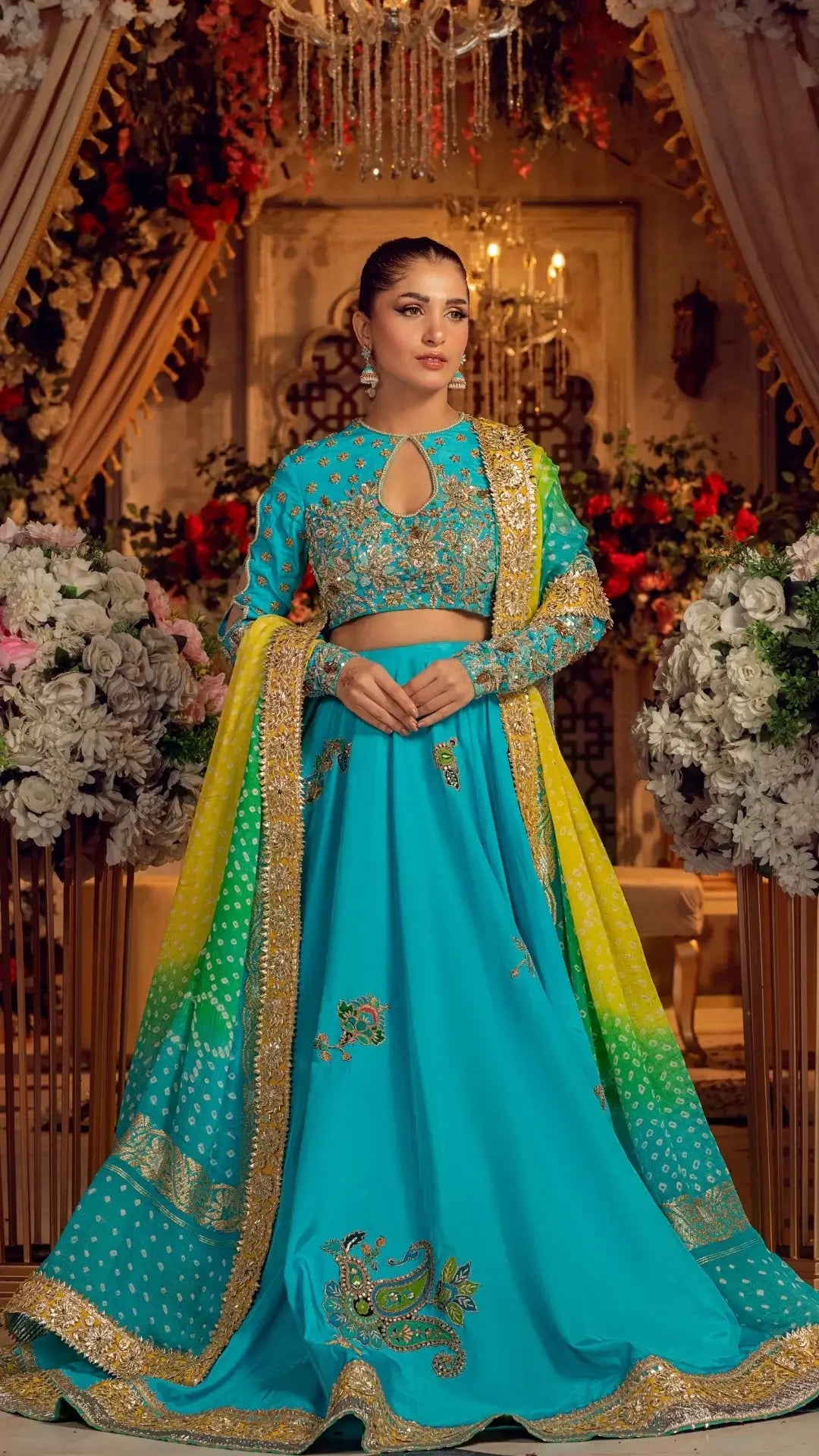 Woman in a traditional turquoise and yellow outfit with floral decorations in an elegant indoor setting.